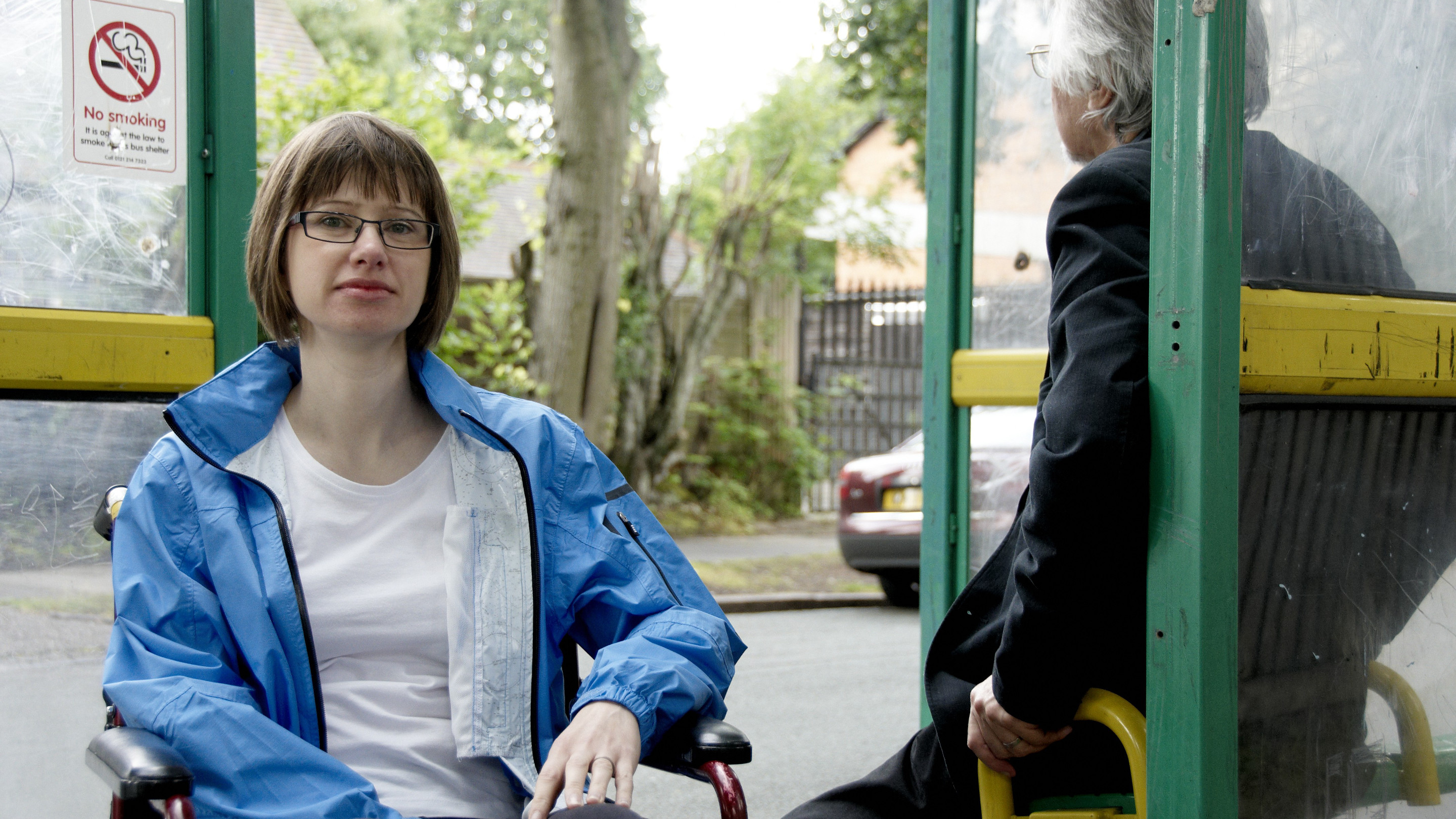 Woman in wheelchair and man sat on bench at bus stop