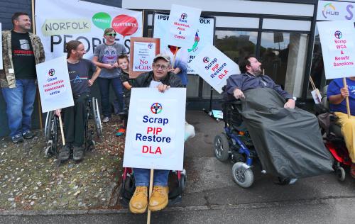 Image of disabled people protesting in Norwich. A man holds a banner which says 'Scrap PIP. Restore DLA'